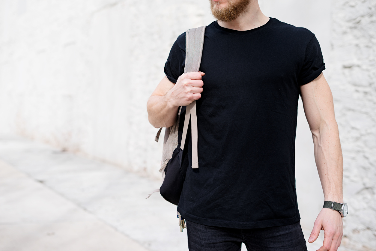 Young muscular bearded man wearing black tshirt and backpack posing outside. Empty white concrete wall on the background. Hotizontal mockup, front view.